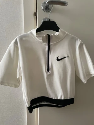 Vit croppad Nike topp med halv zip - Snygg vit croppad topp från Nike med svart logga på bröstet och svart kant nertill. Toppen har korta ärmar och en half zip-dragkedja framtill som ger en sportig vibe. Perfekt till jeans eller träningsbyxor.