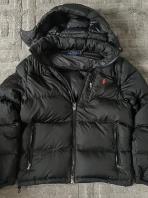 Ralph Lauren jacka - Snygg svart dunjacka från Polo Ralph Lauren med avtagbar huva och två stora dragkedjefickor framtill. Jackan har en röd broderad logga på bröstet och stilren design som ger en puffig look. Perfekt för kalla vinterdagar och riktigt varm. (ÄKTA)