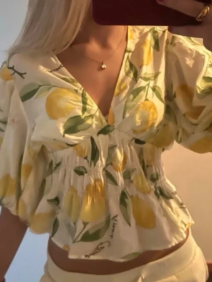 Blommig croppad blus med citroner - Söt croppad blus med citronmönster i gult och grönt på vit botten. Blusen har v-ringning, volangdetaljer och puffiga korta ärmar. Perfekt för sommarens alla tillfällen.