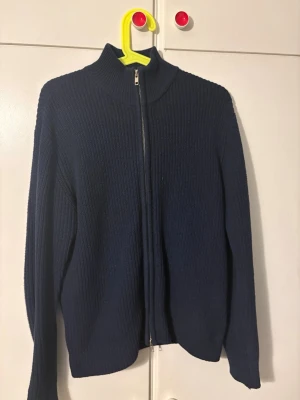 Mörkblå stickad ziptröja - Mörkblå stickad tröja med dragkedja framtill och hög krage. Tröjan har lång ärm och ribbstickad struktur, perfekt för lager-på-lager-stil. Enkel och stilren design som passar till mycket.