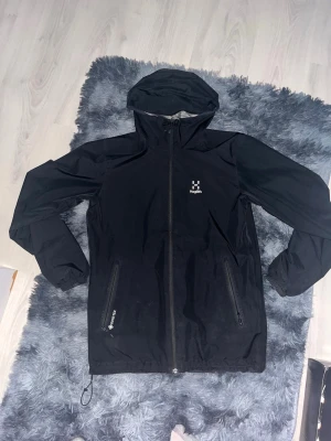 Svart regnjacka från Haglöfs Gore-Tex - Svart regnjacka från Haglöfs med Gore-Tex-material som håller dig torr. Jackan har huva, hel dragkedja framtill, två sidofickor med dragkedja och meshfoder på insidan för extra ventilation. Perfekt för regniga dagar och outdoor-äventyr.