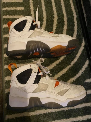 Orange Jordan jumpman - Snygga high top sneakers i vitt med detaljer i orange och svart. Skorna har robust sula i grått och vitt, meshpaneler för extra ventilation och klassisk snörning. Perfekta för dig som gillar sportig och retro stil. Materialmix av läder och textil. 