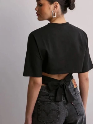 Svart croppad topp med knyt i ryggen - Trendig svart croppad topp med korta ärmar och snygg knytning i ryggen. Toppen har en tight passform och är perfekt för dig som vill ha en stilren men ändå unik look. Tillverkad i mjukt bomullsmaterial som känns skönt mot huden.