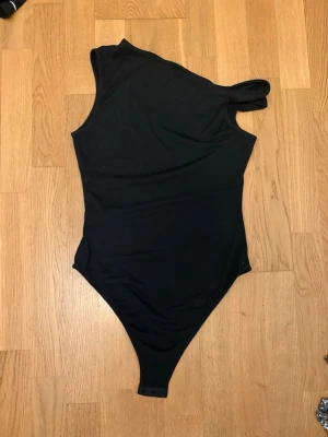 Svart one shoulder body - Snygg svart body med one shoulder-design och asymmetrisk axel. Tillverkad i stretchigt material som sitter tight mot kroppen. Perfekt att styla med jeans eller kjol för en trendig look. Knäppning i grenen för enkel på- och avtagning.