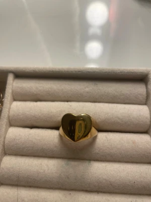  hjärtformad ring - Trendig ring i guldfärgad metall med en blank, hjärtformad front. Ringen har en chunky design som verkligen sticker ut och passar perfekt för dig som gillar statement-smycken. Perfekt att matcha med andra accessoarer för en personlig stil. Säljer för att den inte passade så har visat exempel på storlek på en bild. Inte riktigt guld