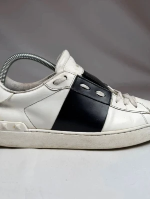 Valentino opens - Snygga sneakers från  Valentino i vitt läder med en bred svart panel på sidan. Klassisk låg modell med vita skosnören och diskreta detaljer. Perfekta för dig som gillar minimalistisk och stilren design.