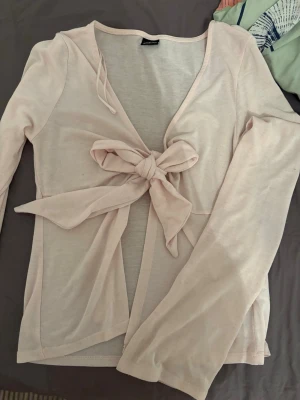 Ljusrosa cardigan med knytband - Supersöt ljusrosa cardigan med långa ärmar och ett stort knytband framtill. Tunn och mjuk i materialet, perfekt att slänga över ett linne eller topp. Öppen framtill och ger en avslappnad men ändå chic look. 