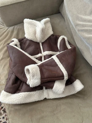Brun teddyjacka med vit fuskpäls - Säljer en riktigt snygg brun teddyjacka med vit fuskpäls på krage, ärmslut och nederkant. Jackan har en hög krage, två fickor och dragkedja framtill. Perfekt för dig som vill ha en varm och trendig look i vinter.