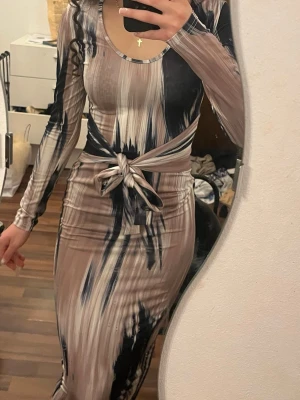 Mönstrad långklänning med knyt - Säljer en långärmad långklänning med abstrakt mönster i beige, svart och grå toner. Klänningen har en tight passform och ett snyggt knyt i midjan som framhäver siluetten. Materialet känns stretchigt och mjukt, perfekt för dig som gillar en kroppsnära look.