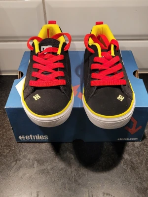 Svarta Etnies DC sneakers - Snygga svarta DC sneakers med stora vita DC-loggor på sidan, röda skosnören och gul insida. Skorna har lågt skaft och vit sula med gul kant. Perfekta för barnet som gillar streetstyle och vill sticka ut med färgglada detaljer. Oanvända.