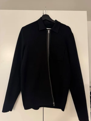 Svart overshirt från Samsøe Samsøe - Stilren svart overshirt från Samsøe Samsøe med rak passform, klassisk krage och sned dragkedja framtill. En bröstficka på vänster sida. Perfekt att bära som lager över en t-shirt. Materialet känns mjukt och lite tjockare.