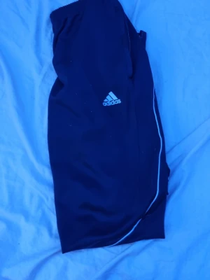 Svarta träningsbyxor från Adidas - Svarta träningsbyxor från Adidas med vitt tryckt logotyp på låret och vita detaljer längs benen. Byxorna har resår i midjan och är tillverkade i lätt och funktionellt material, perfekt för sport och träning.