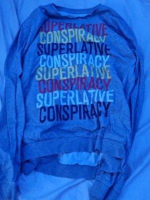Grå sweatshirt från WeSC - Grå sweatshirt från WeSC med tryck i flera färger på bröstet där det står 'SUPERLATIVE CONSPIRACY' i olika stilar. Tröjan har rund halsringning och ribbade muddar vid ärmslut och nederkant. Perfekt för en avslappnad streetwear-look.