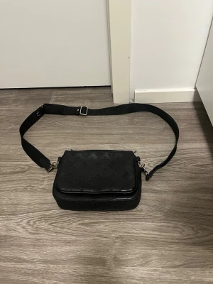 Svart axelväska - Säljer en svart axelväska med justerbar axelrem och klassiskt monogrammönster i läder. Väskan har en fyrkantig form och silverfärgade detaljer. Perfekt för dig som vill ha en stilren och exklusiv accessoar.