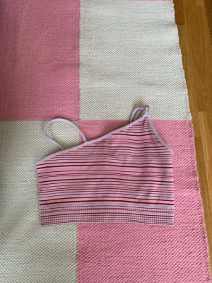 Lila och röd randig crop top - Säljer en ribbad crop top med asymmetrisk design och tunna axelband. Toppen är lila med röda och rosa ränder, perfekt för träning eller sommarens aktiviteter. Materialet är stretchigt och mjukt för en bekväm passform.