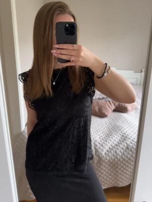 Svart spetsblus med kort ärm - Snygg svart blus i spets med fina detaljer och korta ärmar. Blusen har ett feminint mönster och vågig kant vid ärmslut och nederkant. Perfekt att styla med både kjol och jeans för en trendig look.