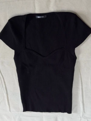 Svart ribbad topp från Gina Tricot - Snygg svart ribbad topp från Gina Tricot med korta ärmar och markerad sweetheart-urringning. Toppen har en figurnära passform och är tillverkad i stretchigt material som sitter bekvämt. Perfekt att styla med jeans eller kjol.