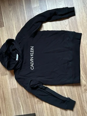 Svart hoodie från Calvin Klein - Svart hoodie från Calvin Klein med stor vit logga på bröstet. Klassisk modell med huva och långa ärmar. Tillverkad i mjukt bomullsmaterial som känns skönt mot huden. Perfekt för en avslappnad och stilren look. Nypris 799, inköpt på zalando.