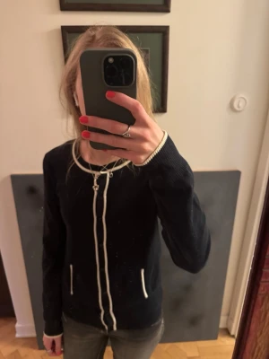 Marinblå kofta med vita detaljer - Snygg marinblå/svart kofta med vita kontrastkanter längs knäppningen, halsringningen och fickorna. Koftan har långa ärmar, rund hals och knäpps med svarta knappar framtill. Perfekt att slänga över en t-shirt eller blus för en stilren look.