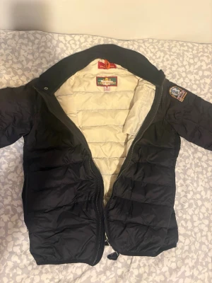 Svart dunjacka från Parajumpers - Snygg svart dunjacka från Parajumpers med beige innerfoder och klassisk patch på ärmen. Jackan har dragkedja framtill, huva och quiltad design som håller värmen. Perfekt för kalla vinterdagar och riktigt stilren look. Hör av dig om du har några frågor. Priset är inte hugget i sten