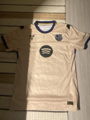 Beige FC Barcelona matchtröja - Säljer en beige FC Barcelona matchtröja med svarta detaljer och blå kant på kragen. Tröjan har klubbmärke, Nike-logga och Spotify som sponsor framtill. Tillverkad i lätt och ventilerande material, perfekt för fotboll eller träning.