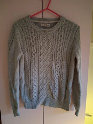 Ljusgrå kabelstickad tröja - Snygg ljusgrå kabelstickad tröja från Ranch Sport med klassiskt flätmönster och rund halsringning. Tröjan har långa ärmar och ribbade muddar vid ärmslut och nederkant. Perfekt för lager-på-lager och en stilren look.