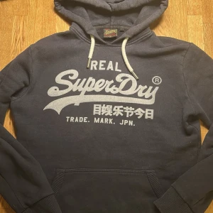Mörkblå Superdry hoodie - Säljer en mörkblå hoodie från Superdry med stor vit logga och text på bröstet. Hoodien har huva med vita dragsnören och en klassisk magficka framtill. Perfekt för dig som gillar streetwear och vill ha något bekvämt och snyggt.