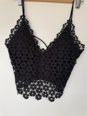 Svart spetsig croppad linnetopp - Svart croppad linnetopp med blommig spets och tunna axelband. Toppen har v-ringning och dekorativa blommor längs kanterna, samt en öppen rygg med smala band. Perfekt för en trendig och stilren look.