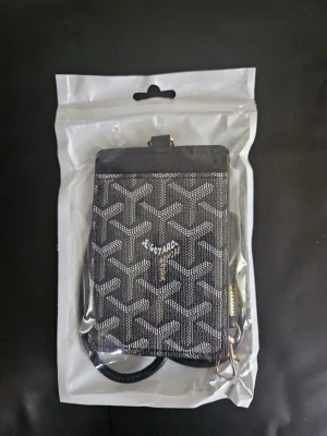 Svart korthållare med snodd - Snygg svart korthållare med transparent fönster på framsidan och matchande svart snodd med metallspänne. Perfekt för att bära ID eller busskort runt halsen. Korthållaren har ett diskret mönster och är gjord i konstläder.