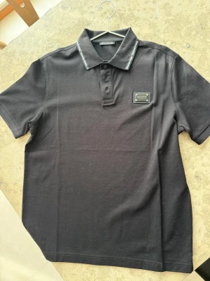 Svart pikétröja från Balmain - Snygg svart pikétröja från Balmain med klassisk krage och knappslå. Tröjan har en exklusiv metallbricka med Balmain Paris på bröstet och diskreta detaljer på kragen. Perfekt för dig som vill ha en stilren och lyxig look.