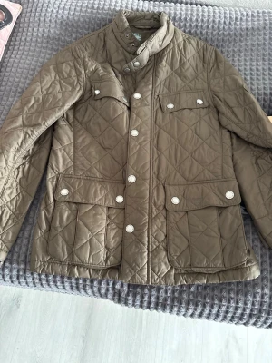 Quiltad grön jacka från Barbour - Snygg quiltad jacka från Barbour i brun färg med vita knappar och hög krage. Jackan har flera fickor framtill och klassiskt rutmönster i materialet. Perfekt för dig som gillar stilren och tidlös design.