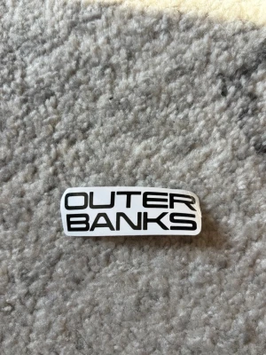 Outer Banks klistermärke svartvit - Snyggt klistermärke med texten 'OUTER BANKS' i svart på vit bakgrund. Rektangulär form med rundade hörn, perfekt att sätta på dator, mobil eller vattenflaska. Gjort i plastmaterial.
