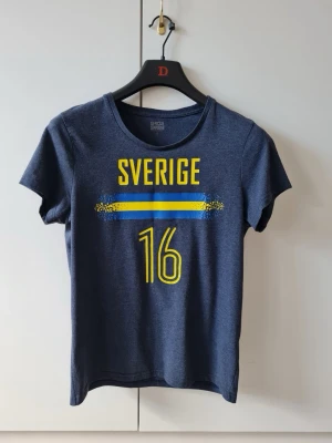 Blå Sverige t-shirt med nummer 16 från Stadium strl S - Mörkblå t-shirt från Stadium med tryck i gult och blått: 'Sverige' och siffran 16 på framsidan från Stadium. Gjord i 100% bomull och har en klassisk rund hals och korta ärmar. Storlek S. Kan hämtas i Vallentuna.