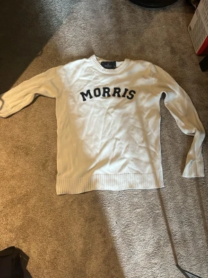 Vit stickad tröja från Morris - Vit stickad tröja från Morris med svart text på bröstet. Tröjan har rund halsringning och ribbstickade muddar vid ärmslut och nederkant. Perfekt för en stilren och klassisk look.