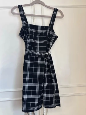 Svartvit rutig klänning med bälte - Stilren svart klänning med vitt rutmönster och breda axelband. Klänningen har en rak passform och medföljande bälte i midjan för extra form. Perfekt för dig som gillar klassiska mönster och enkel design. Super snygg till sommaren! 💕