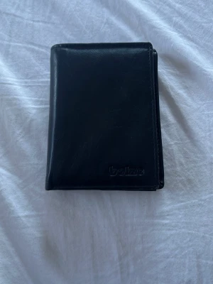 Svart läderplånbok från Bolzo - Stilren svart plånbok i äkta läder från Bolzo. Plånboken har flera kortfack på båda sidor och en klassisk vikbar design. Perfekt för dig som vill ha en enkel och snygg accessoar med plats för många kort. Plånboken är i använt skick men fortfarande väldigt fin.