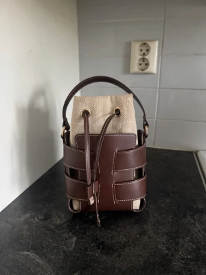Brun bucket bag med canvasdetaljer - Säljer en snygg bucket bag med brunt läder och beige canvasinsats. Band följer med. Väskan har jag fått i present och vet inte vart den är köpt från. 