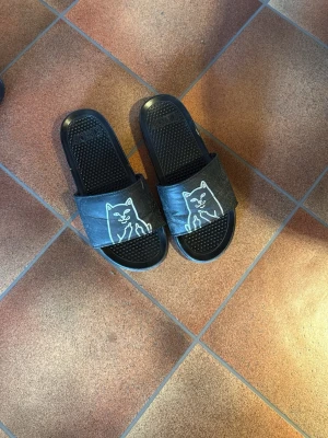 Tofflor  - Säljer ett par svarta slides från RIPNDIP med ikoniskt vitt kattmotiv på remmen. Sandalerna har en klassisk slip-on design och är tillverkade i syntetmaterial med räfflad sula för extra grepp. Perfekta för chill dagar eller att glida runt i hemma.