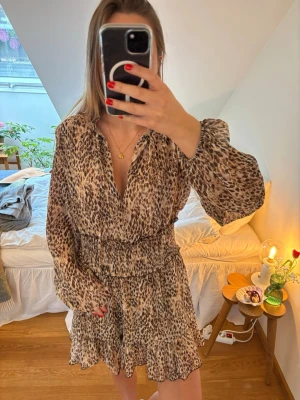 Leopardmönstrad klänning - Kort leopardmönstrad klänning med långärmad design och v-ringning. Klänningen har smock i midjan för en snygg passform och volangdetaljer längst ner. Tillverkad i ett lätt, luftigt material som ger en skön känsla.