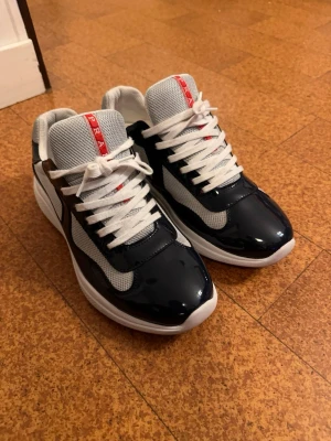 Prada sneakers i svart och vitt - Snygga Prada sneakers i mörk blåa med röda detaljer och vit sula. Ovandelen är i en mix av glansigt läder och mesh-tyg för en sportig look. Klassisk låg modell med vita skosnören och tydlig Prada-logga på plös och häl.