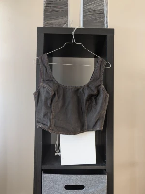 Svart denim korsettopp - Trendig svart korsettopp i denim med breda axelband och markerade sömmar. Toppen har en croppad passform och dragkedja i ryggen för enkel påklädning. Perfekt att styla med höga jeans eller kjol för en edgy look.
