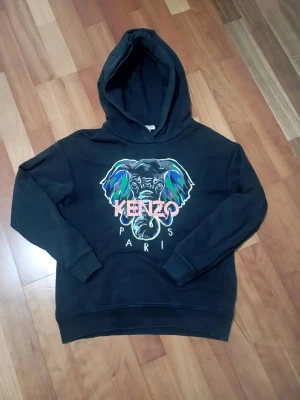 Kenzo svart hoodie med elefanttryck - Svart hoodie från Kenzo Kids med färgstarkt elefantmotiv och texten 'Kenzo Paris' på bröstet. Hoodien har huva, ribbade muddar och är tillverkad i mjukt material som känns skönt mot huden. Perfekt för dig som gillar streetstyle och vill ha något unikt.