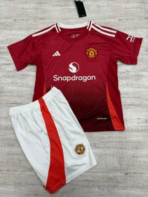 Manchester United matchställ Adidas - Säljer ett Manchester United matchställ med röd kortärmad tröja och vita shorts. Tröjan har klubbmärke, Adidas-logga och Snapdragon-tryck, samt vita ränder på axlarna. Shortsen har röd sidopanel och klubbmärke. Materialet är lätt och andas, perfekt för träning.