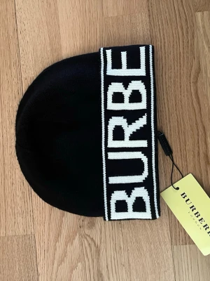 Svart Burberry mössa med logotyp - Cool svart mössa från Burberry med stor vit logotyp på uppviket. Tillverkad i mjukt stickat material som håller dig varm och ger en snygg look. Perfekt för dig som vill ha en trendig accessoar med exklusiv känsla.