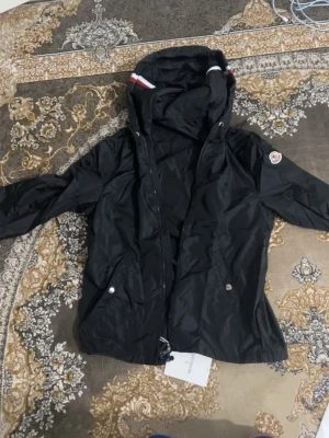 Svart vindjacka från Moncler - Snygg svart vindjacka från Moncler med huva och dragkedja framtill. Jackan har Moncler-logga på ärmen, silverfärgade knappar vid fickorna och detaljer i rött och vitt på huvan. Perfekt för dig som vill ha en stilren och sportig look.