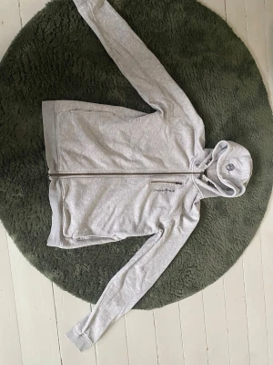 Ljusgrå hoodie från Sail Racing - Ljusgrå hoodie från Sail Racing med dragkedja framtill och broderad logga på bröstet. Utrustad med huva och två sidofickor. Insidan är fodrad med mjuk teddy för extra komfort. Perfekt för chill dagar eller när du vill ha en sportig look.