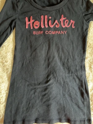 Navy blå långärmad topp från Hollister - Navy blå långärmad topp från Hollister med broderad röd text 'Hollister Surf Company' framtill. Jättebra skick förutom att den är lite nopprig och har några enstaka lösa trådar. Storlek Xs.