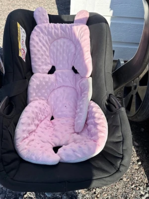 Rosa sittdyna med öron till barnvagn - Säljer en supermjuk och vadderad rosa sittdyna med söta öron, perfekt för barnvagn eller bilbarnstol. Dynan har små prickar i tyget och är formad för att ge extra stöd och komfort till barnet. Passar de flesta barnvagnar och babyskydd.