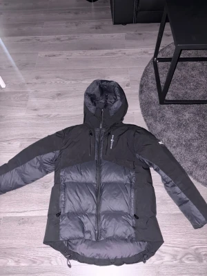 Svart dunjacka från Sail racing - Snygg svart dunjacka från Sail racing med huva och dragkedja framtill. Jackan har flera praktiska fickor, både på bröstet och sidorna, samt justerbar nederkant. Perfekt för kalla vinterdagar och outdoor-äventyr. Ny pris 3500kr