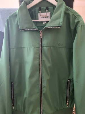 vindjacka från Lacoste - Snygg vindjacka från Lacoste med dragkedja framtill och två fickor med dragkedja. Klassisk krage och diskret Lacoste-logga på bröstet. Jackan är tillverkad i ett lätt och smidigt material som passar perfekt till vår och sommar. Pris kan diskuteras vid snabb affär!!